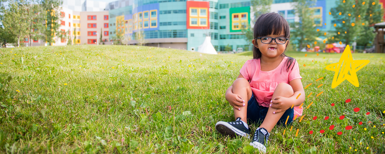 One Child Every Child - a young girl with glasses and a pink shirt sits on the grass in front of the Alberta Children's Hospital
