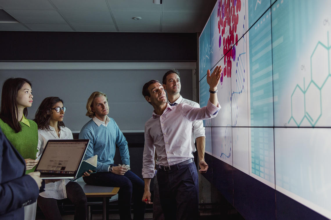 Group of business professionals in a dark room standing in front of a large data display screen with information.