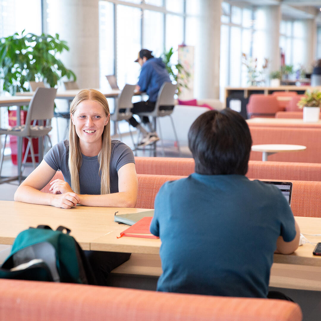 Two people sit at a table in a common area chatting